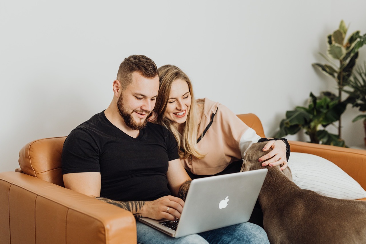a smiling couple and dog looking at a laptop